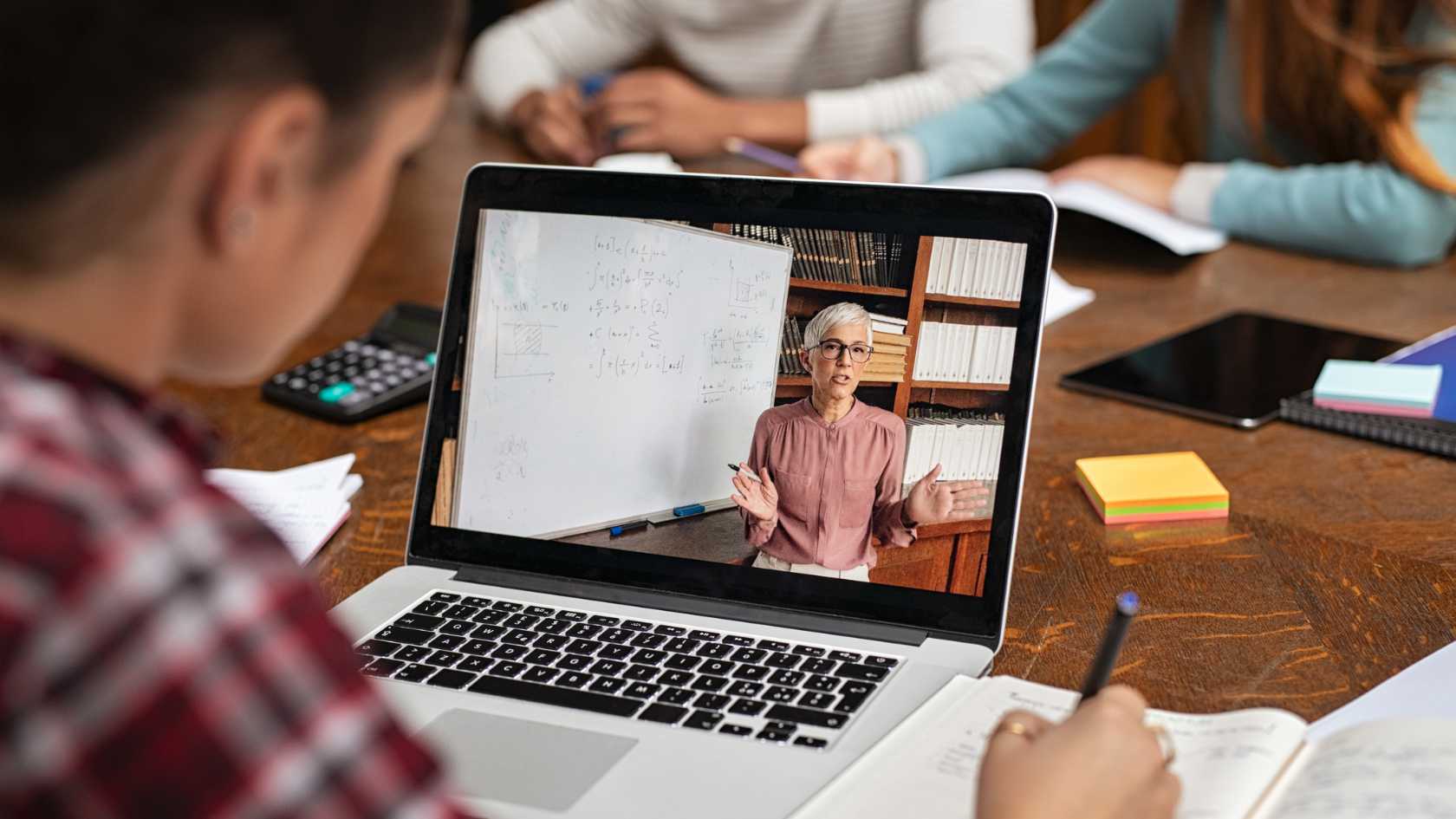 Blick über die Schulter einer Studentin, die in einen Laptop auf dem Tisch vor ihr schaut. Auf dem Laptop ist das Video einer Lehrveranstaltung zu sehen.