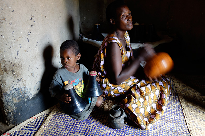 Während Edith in Uganda die Milch mit speziellen Gefässen buttert. Daneben ihr Enkel mit zwei ebyanzi-Holztrinkgefässen und zwischen den beiden das Räucher-Tongefäss ekiicunga.