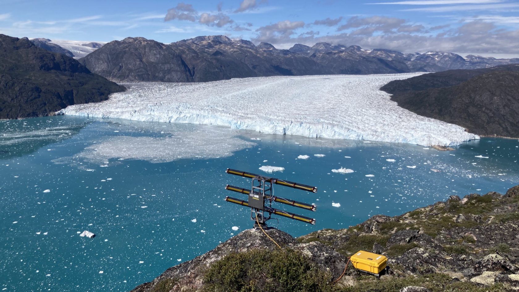 View of the fjord and the three-kilometer-wide calving front of Eqalorutsit Kangilliit Sermiat in southern Greenland. The fiber-optic cable was laid a few hundred meters from the ice wall through the 300-meter-deep water on the seabed. In the foreground is the UZH radar device, which measures calving events and ice movements in order to interpret the data from the fiber-optic cable.