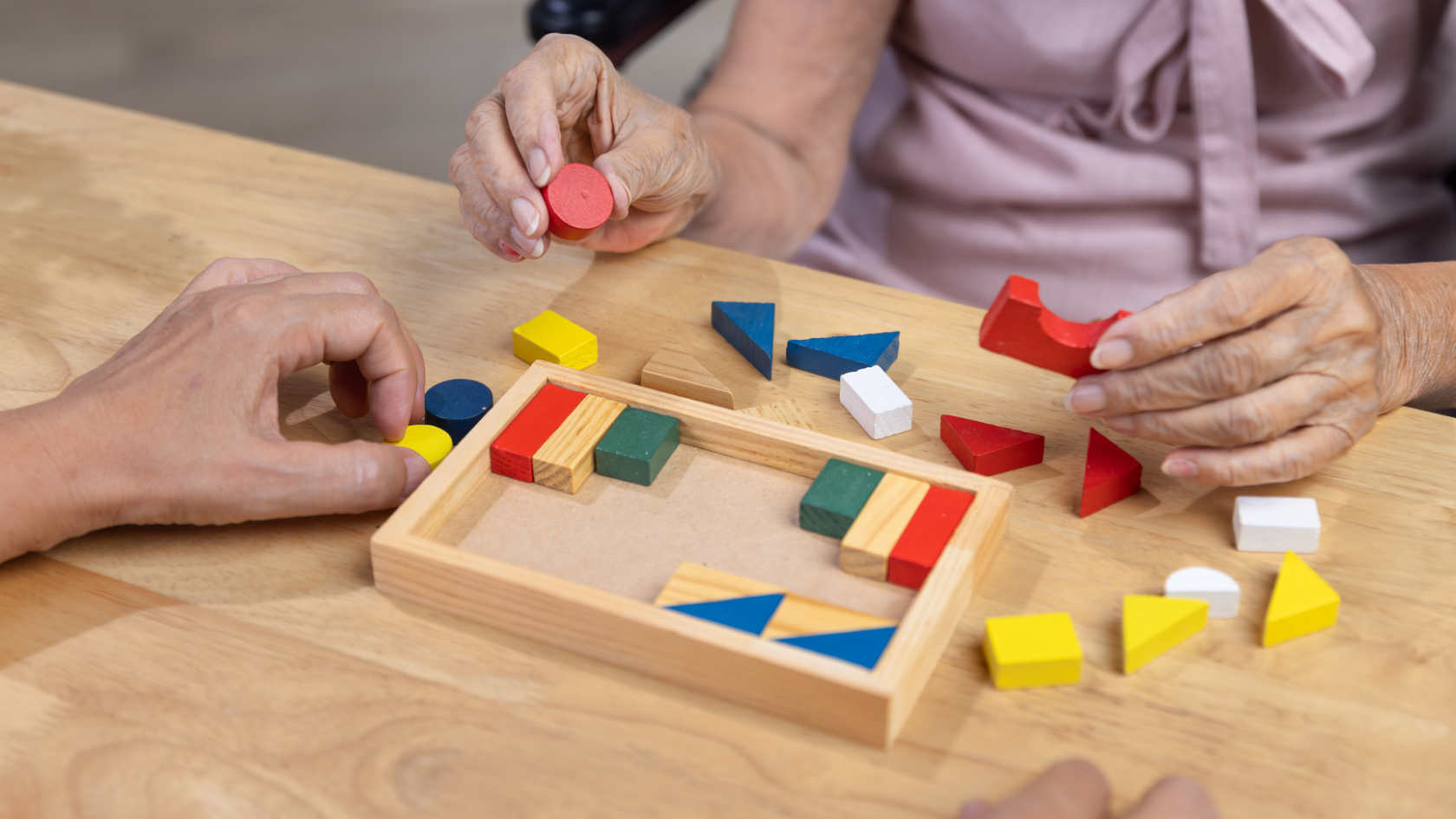 A wooden puzzle and various colored pieces lie on a table, two hands hold puzzle pieces.