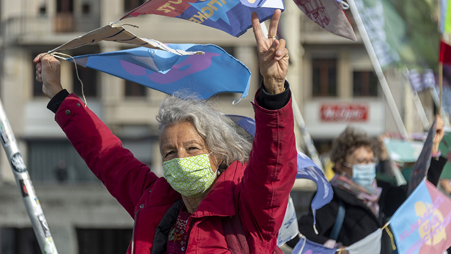 An elderly lady holds posters with one hand, with the other she forms the peace sign.